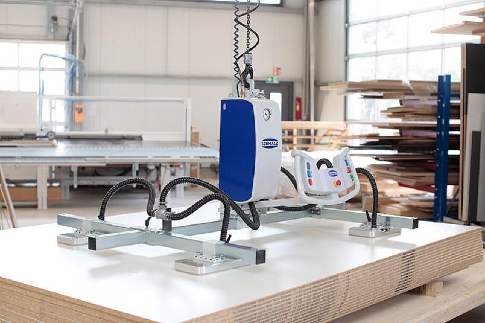 In einer geräumigen, gut beleuchteten Holzwerkstatt mit Bretterstapeln im Hintergrund hängt ein Vakuumheber mit Bedienfeld über großen Sperrholzplatten.