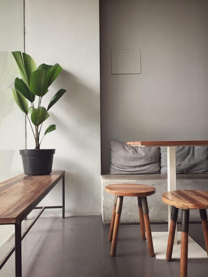 Ein minimalistisches Interieur mit einer Holzbank mit einer Topfpflanze, einem kleinen Holztisch, zwei passenden Hockern und einer Betonbank mit grauen Kissen vor einer hellgrauen Wand.