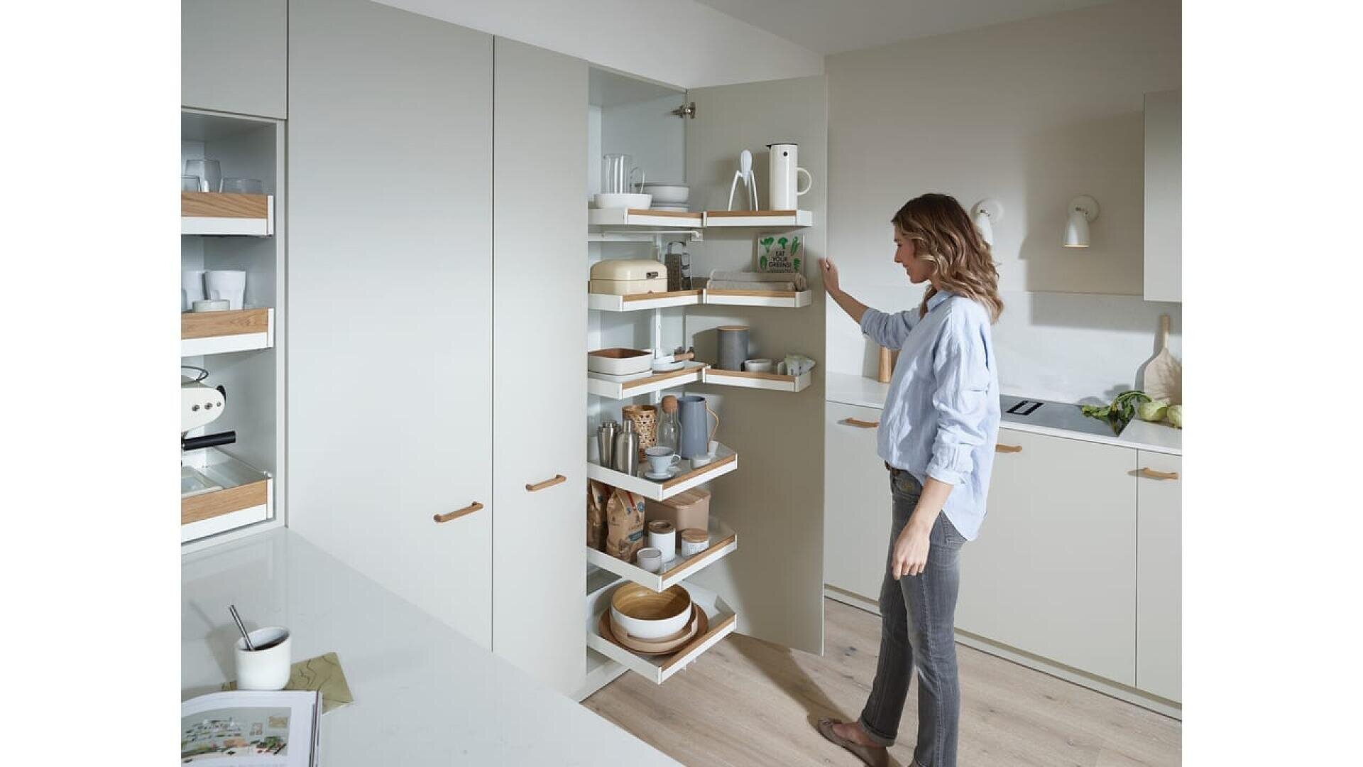 Une femme se tient dans une cuisine moderne, la main tendue vers un meuble d'angle ouvert avec des étagères coulissantes contenant de la vaisselle, des bols et des ustensiles de cuisine. La cuisine est de couleur claire, avec un design minimaliste et des accents en bois.