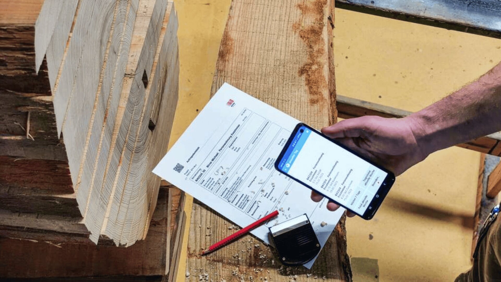 Eine Person hält ein Smartphone, auf dem Daten angezeigt werden, neben einem gedruckten Dokument, einem Bleistift und einem kleinen Gerät auf einem Holzbrett in einer Werkstatt mit Stapeln von geschnittenem Holz.