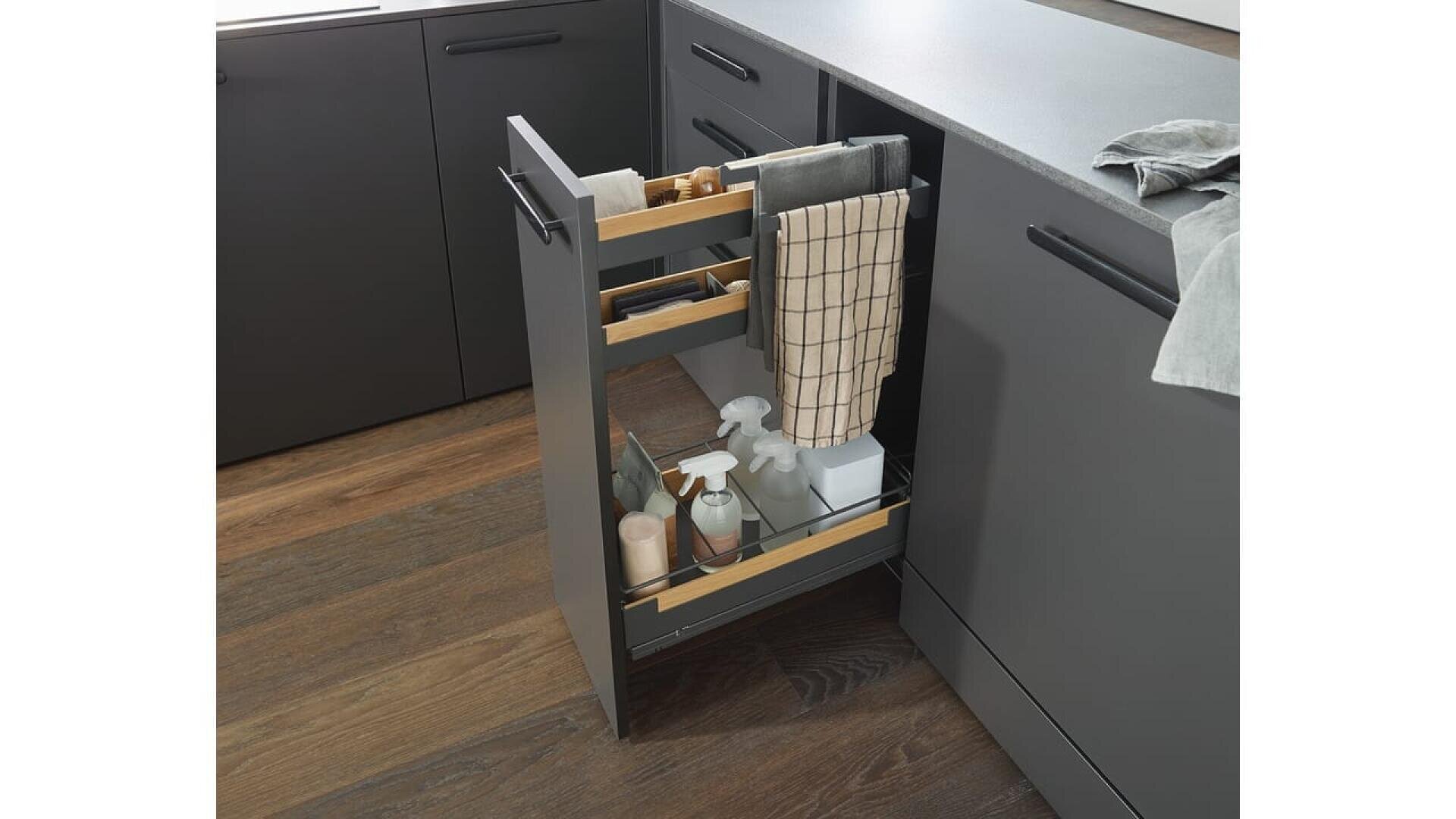 Une armoire de cuisine coulissante permet de ranger les produits de nettoyage, les flacons pulvérisateurs, les éponges et un torchon à carreaux. Les placards sombres et le sol en bois donnent à la cuisine un aspect moderne.