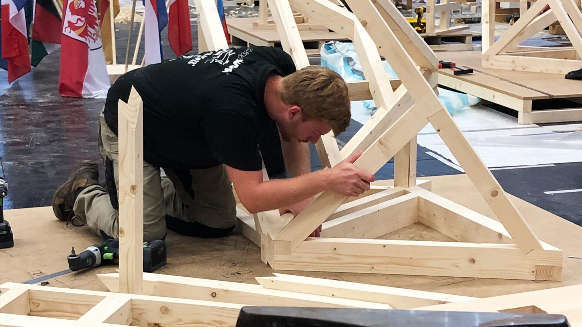 Eine Person kniet auf dem Boden und baut mit Werkzeugen und Holzstücken eine Holzkonstruktion zusammen. Im Hintergrund sind Fahnen zu sehen, die auf einen Wettbewerb oder ein gemeinsames Ereignis hindeuten.