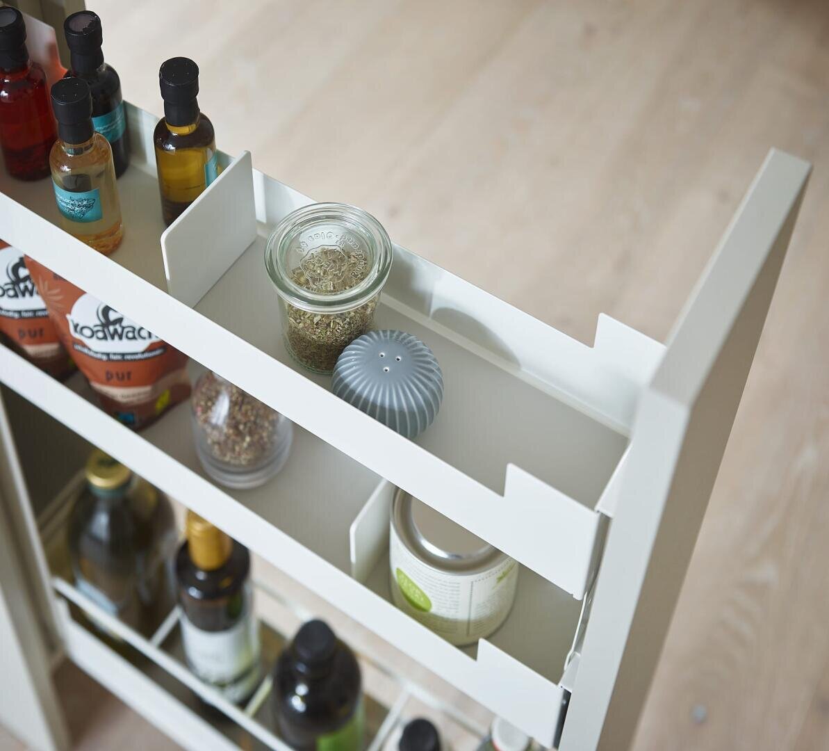 Ein weißes, ausziehbares Küchenregal mit Gewürzdosen, Ölflaschen, Soßenpackungen und einem kleinen runden Behälter auf einem hellen Holzboden.