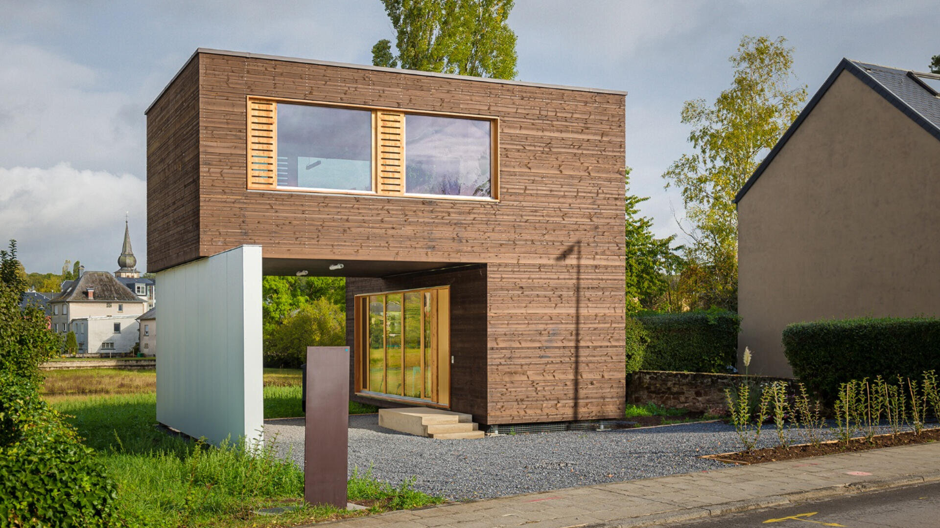 Ein modernes zweistöckiges Haus mit einer Holzfassade, großen Fenstern und einem auskragenden Obergeschoss. Das Haus hat ein minimalistisches Design und ist von Grünflächen und benachbarten Gebäuden umgeben.