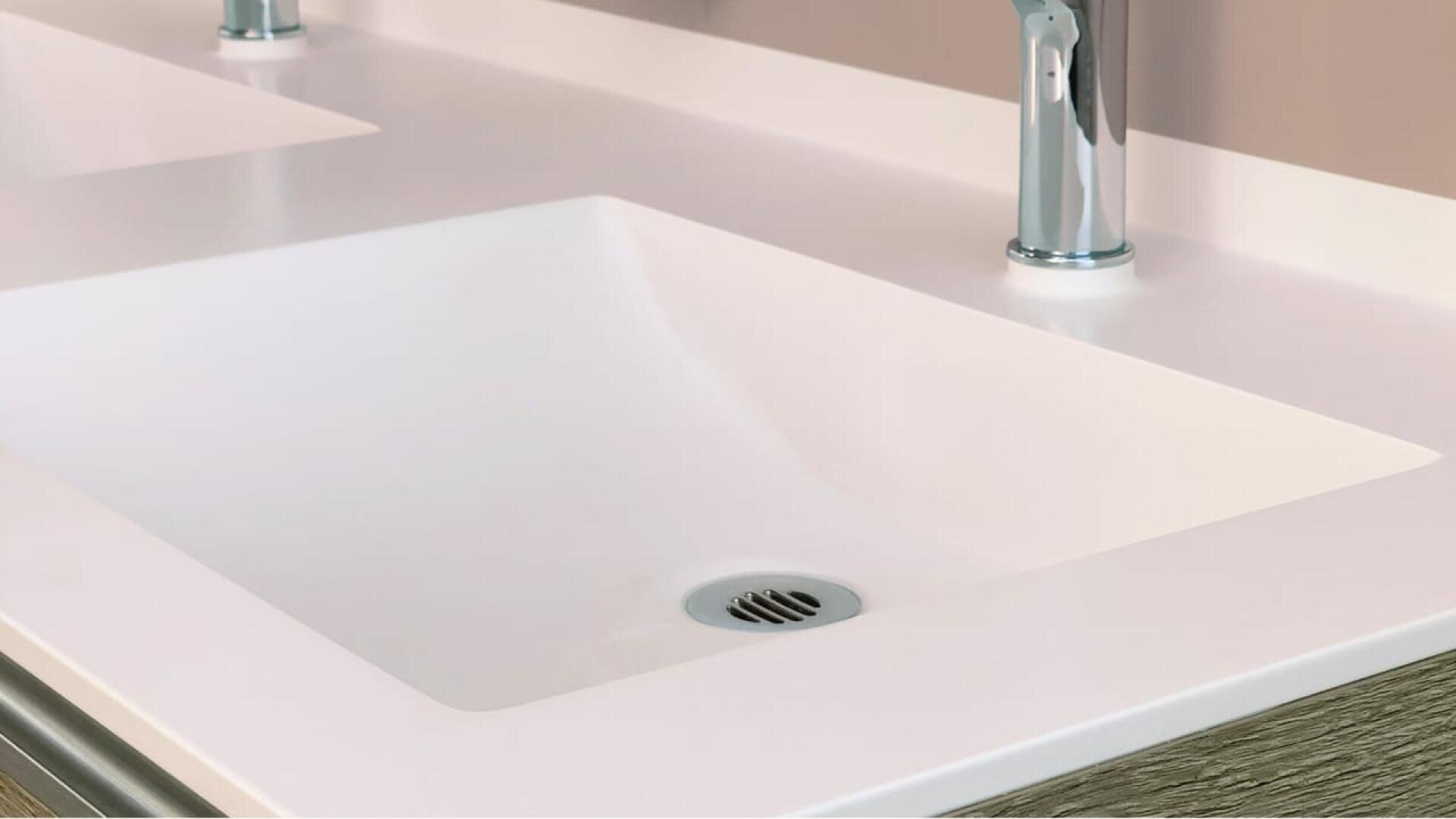 Gros plan sur un évier de salle de bains moderne, blanc et rectangulaire, avec un robinet chromé et un siphon en métal. Le plan de travail est lisse et propre, et une partie d'un autre lavabo identique est visible à côté.