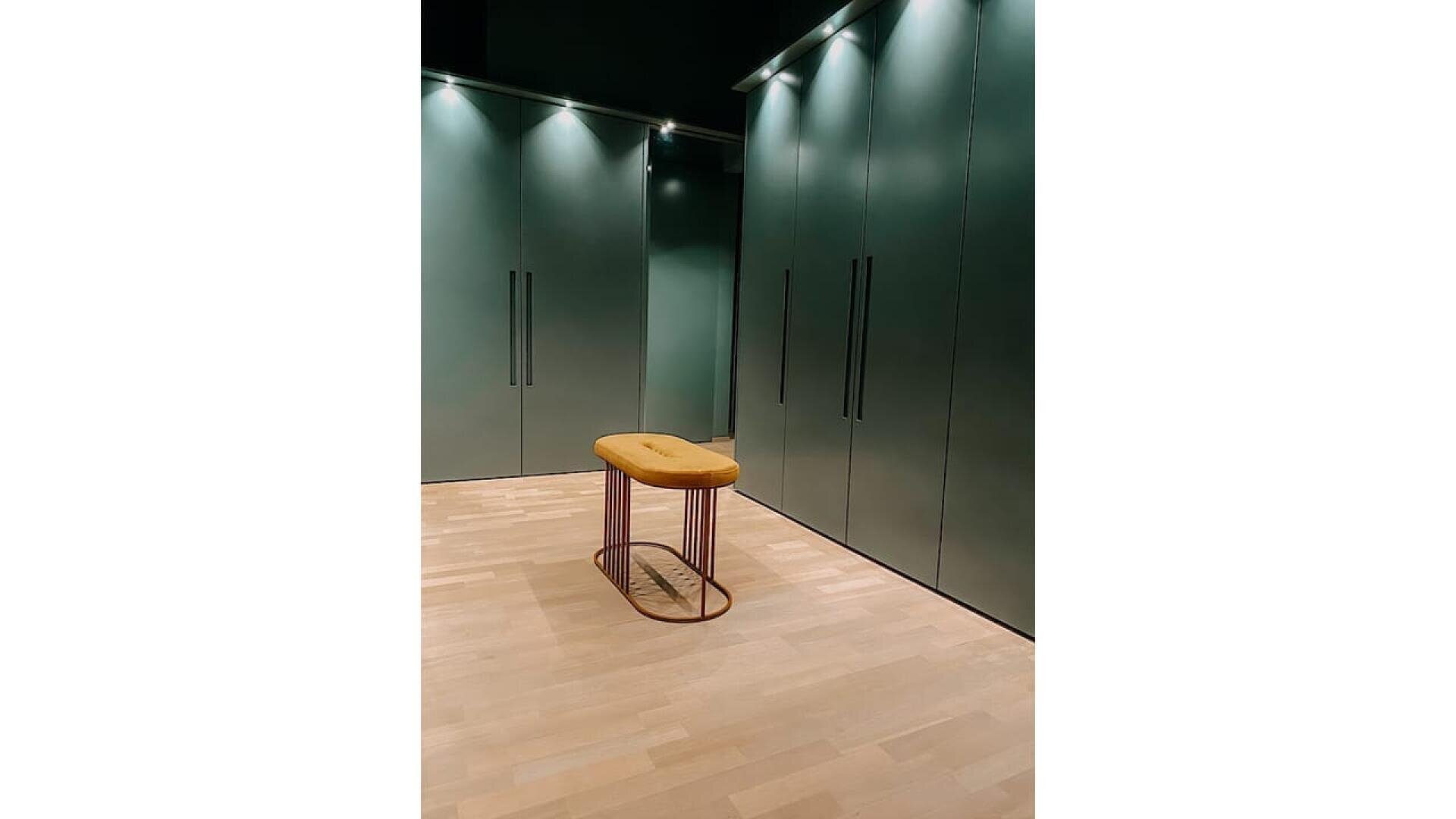 Une petite pièce avec des armoires vertes allant du sol au plafond, un sol en bois clair et un tabouret moderne avec une assise jaune moutarde coussinée et des pieds en métal placés au centre. Des lumières douces au plafond éclairent l'espace.
