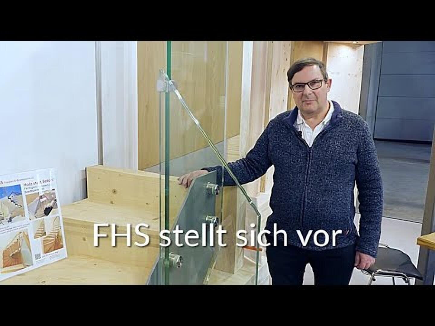 Un homme portant des lunettes et un pull sombre se tient à l'intérieur, à côté d'une structure en verre et d'un présentoir en bois. Le texte sur l'image indique FHS stellt sich vor en allemand.