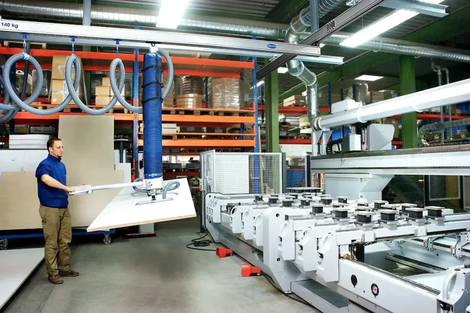 Ein Arbeiter bedient einen Vakuumheber, um eine große Holzplatte in einer modernen industriellen Fabrik mit Materialregalen und modernen Maschinen im Hintergrund zu bewegen.