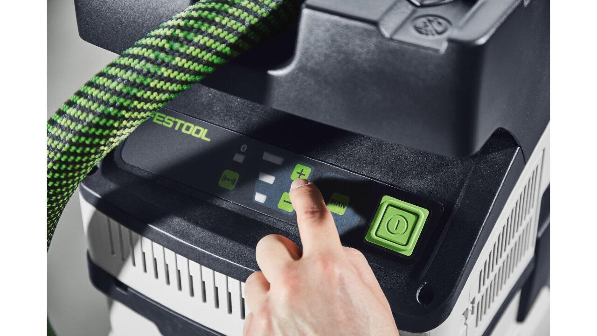 Une main appuie sur le bouton "plus" du panneau de commande numérique d'un outil Festool, un tuyau vert et noir étant visible au-dessus de l'écran.