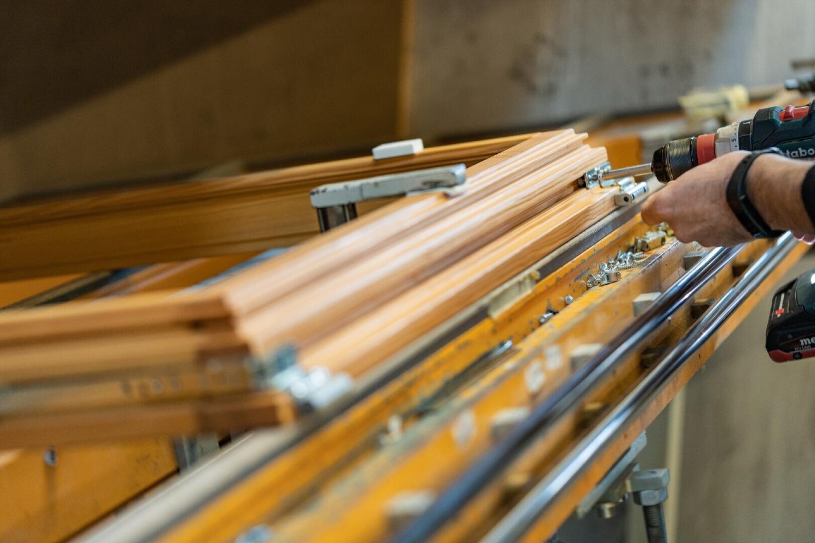 Gros plan sur une personne qui utilise une perceuse pour assembler ou ajuster des cadres ou des poutres en bois dans un atelier ou une usine de fabrication. Sur l'établi, on peut voir différents outils et pièces métalliques.