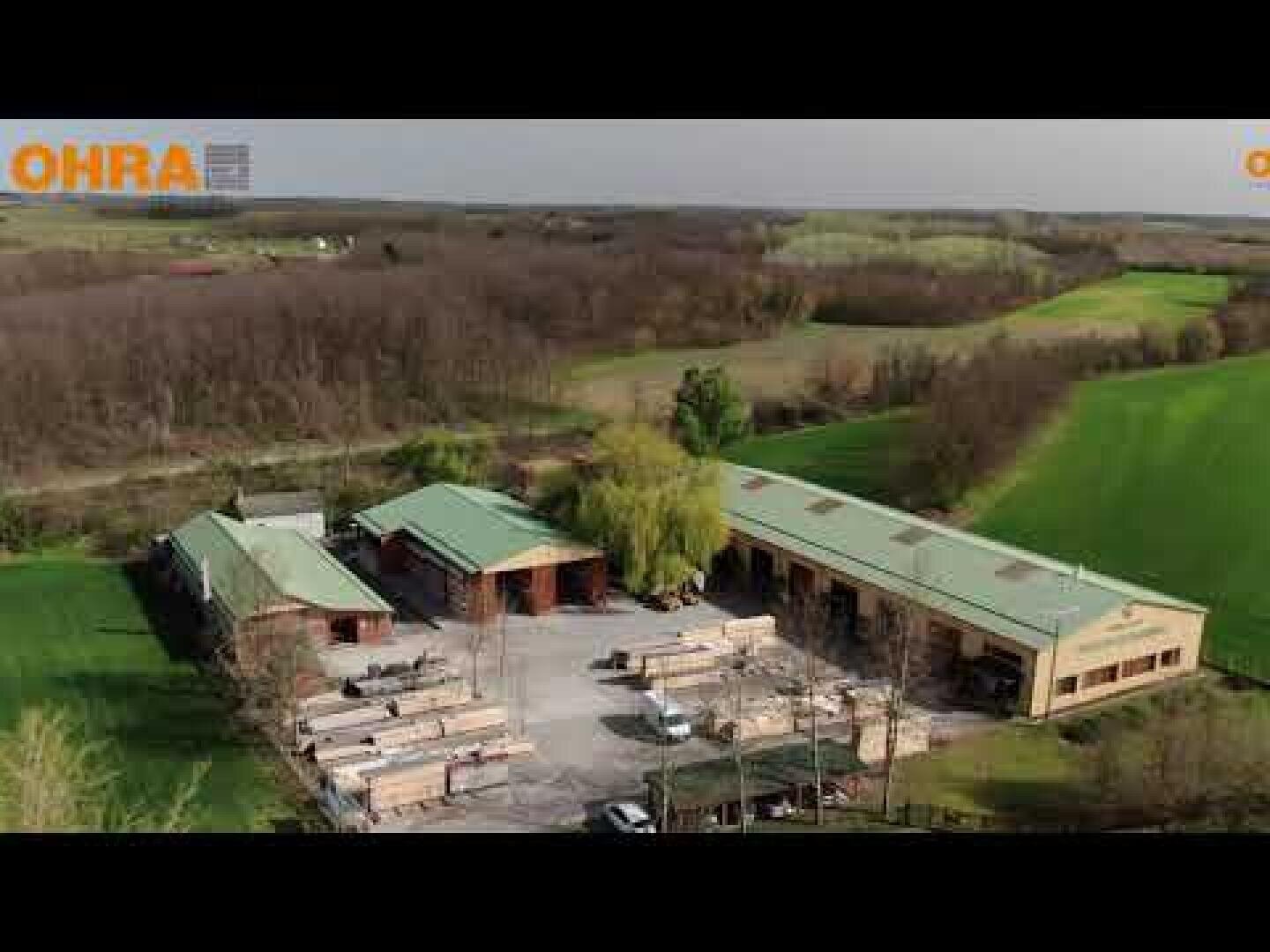 Luftaufnahme einer ländlichen Industrieanlage mit Gebäuden mit grünen Dächern, umgeben von Feldern, Bäumen und sanft geschwungenen Hügeln unter einem teilweise bewölkten Himmel. Das OHRA-Logo ist in der oberen linken Ecke zu sehen.