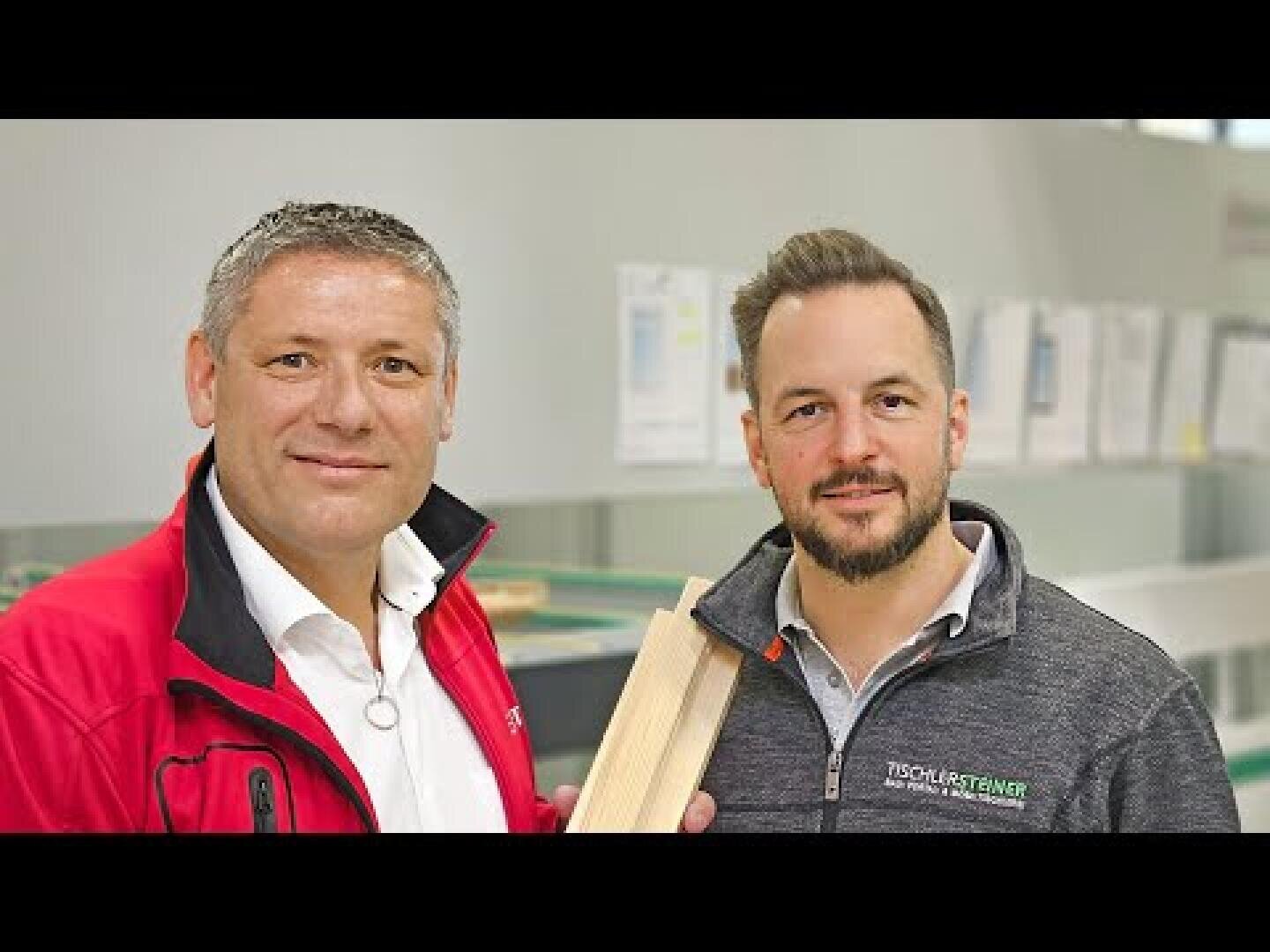 Zwei Männer stehen nebeneinander im Inneren und lächeln beide in die Kamera. Einer von ihnen hält ein Stück Holz in der Hand. Beide tragen legere Geschäftskleidung und Markenjacken. An der Wand im Hintergrund sind Dokumente zu sehen.