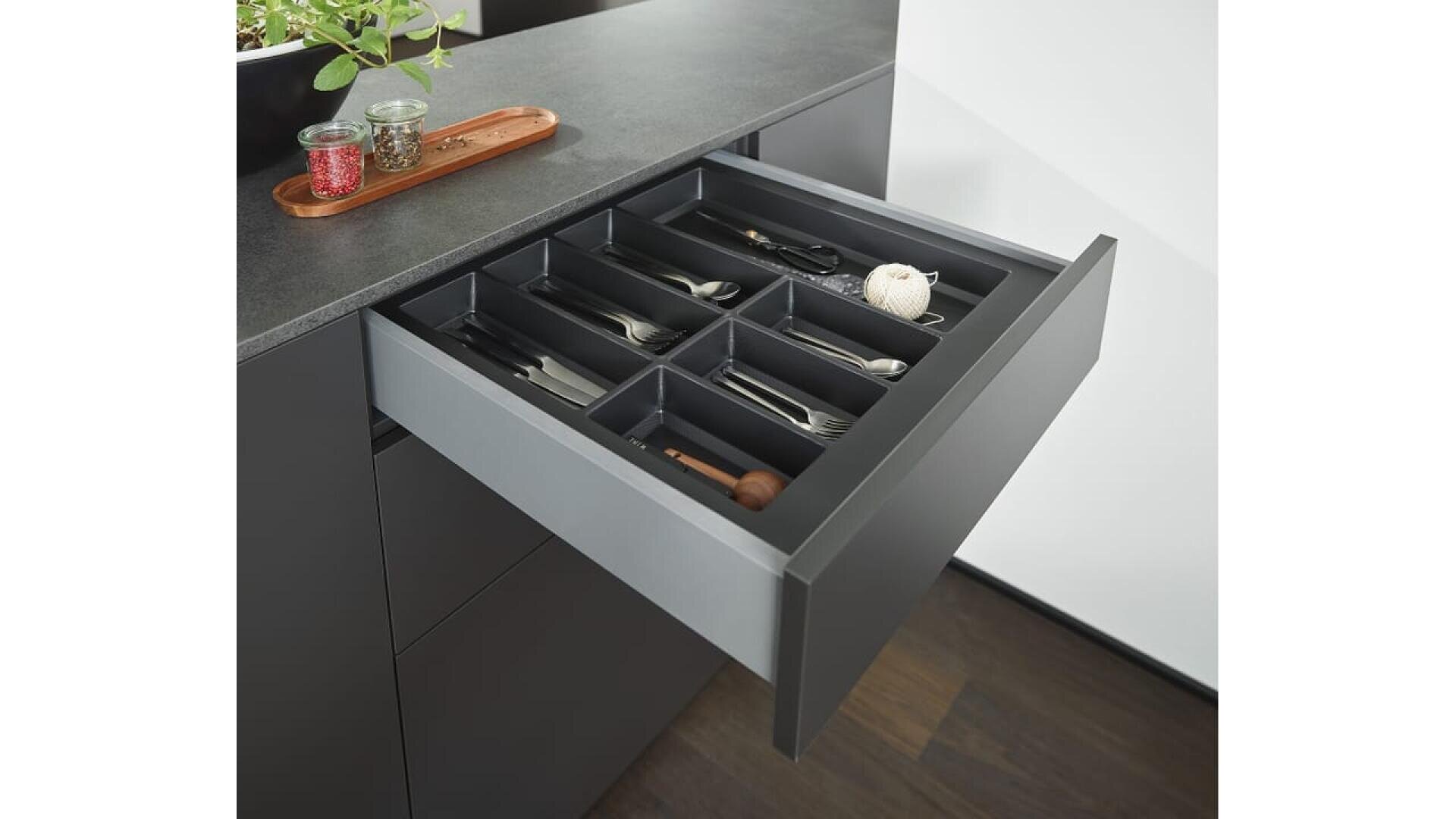 Un tiroir de cuisine moderne est ouvert, montrant un plateau de couverts noir bien rangé avec divers ustensiles, notamment des fourchettes, des couteaux, des cuillères et quelques petits outils de cuisine à l'intérieur de chaque compartiment.