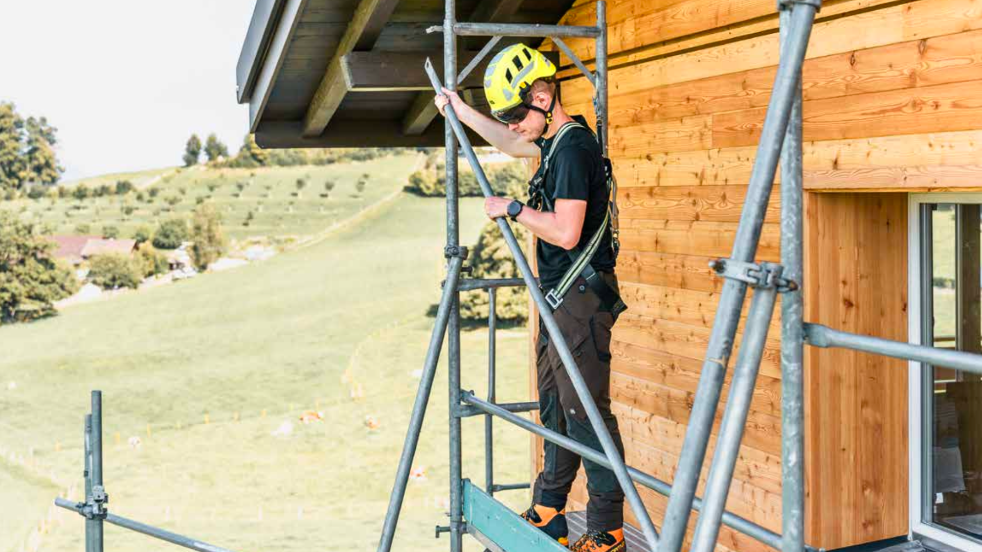 Un ouvrier du bâtiment portant un casque jaune et un équipement de sécurité se tient sur un échafaudage à côté d'un bâtiment en bois, en s'accrochant à un poteau métallique, avec un paysage herbeux et des arbres en arrière-plan.