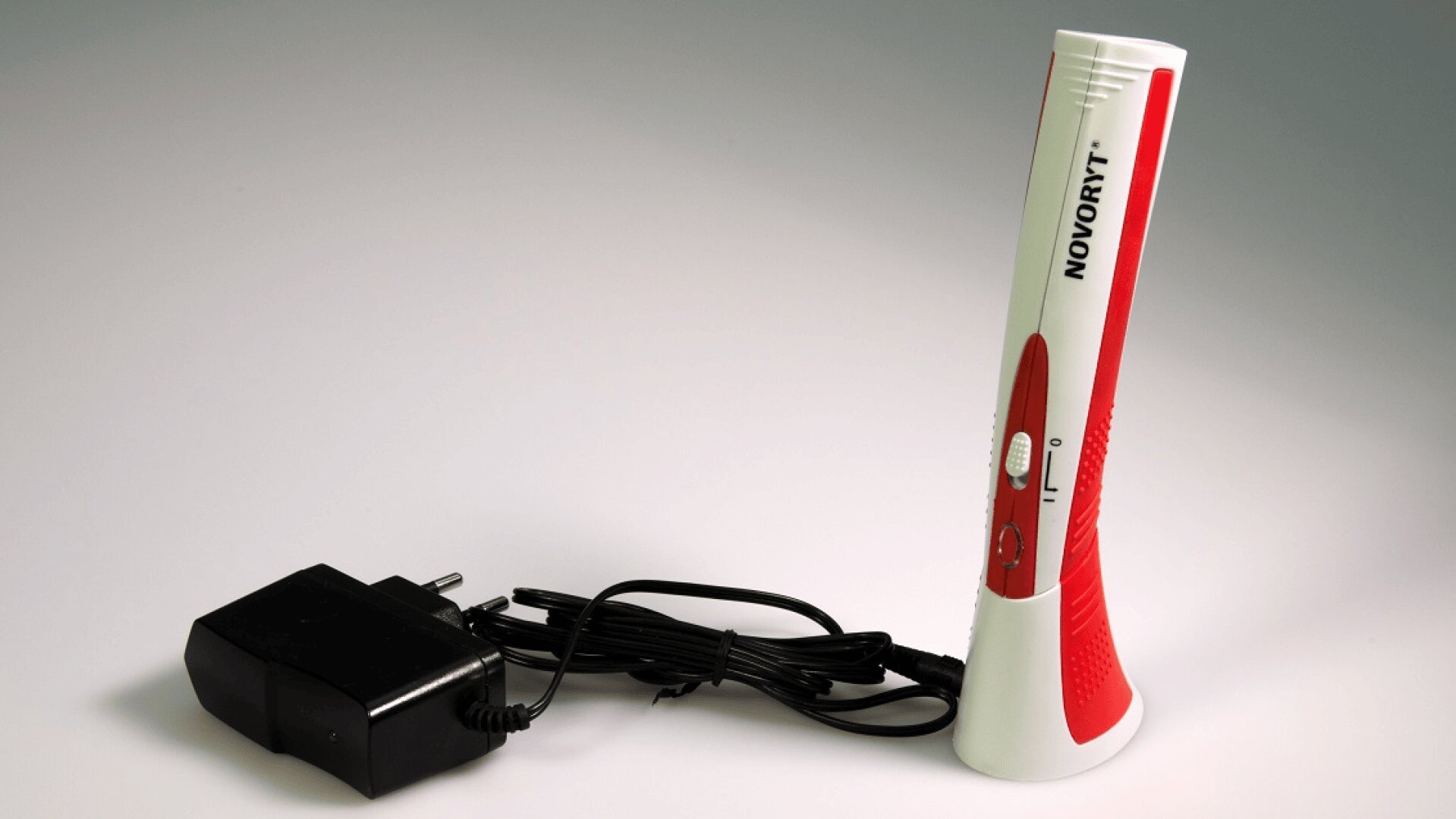Ein rot-weißes elektronisches Handgerät mit der Aufschrift NOVORYT steht aufrecht neben einem schwarzen Netzadapter und einem Kabel auf einer weißen Fläche.