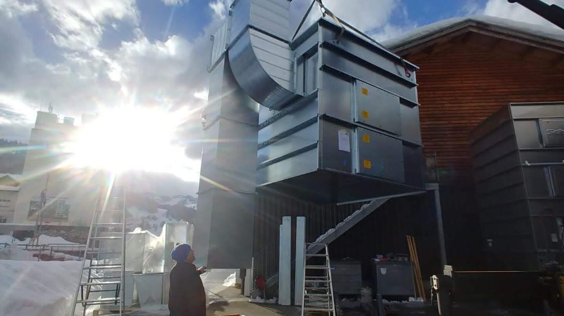 Eine Person steht in der Nähe großer industrieller Metallrohre und Geräte, die im Freien installiert werden, mit Leitern in der Nähe. Schnee bedeckt den Boden und die Gebäude, und die Sonne scheint hell hinter Wolken am Himmel.