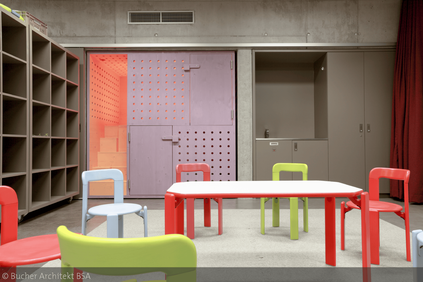 Ein modernes Klassenzimmer mit bunten roten, grünen und blauen Stühlen um einen weißen Tisch. Auf der linken Seite befinden sich Regalfächer, auf der rechten Seite ein Schrank und ein Waschbecken und auf der Rückseite eine lila Tür mit perforierten Paneelen.