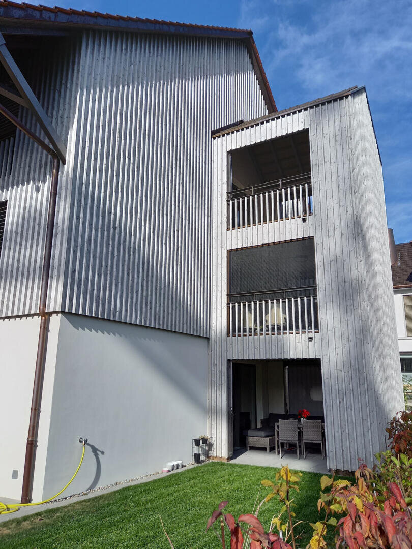 Ein modernes, dreistöckiges Gebäude mit vertikalen Holzlatten an der Außenseite, offenen Balkonen und einer Terrasse im Erdgeschoss mit Gartenmöbeln, neben einem grünen Rasen unter blauem Himmel.