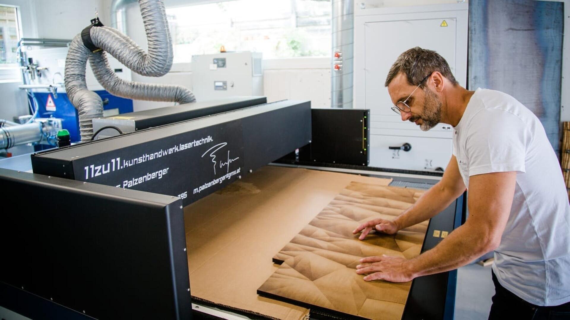 Dans un atelier moderne, un homme portant des lunettes et un t-shirt blanc inspecte un morceau de bois sculpté de manière complexe qui sort d'une grande machine industrielle de découpe au laser.