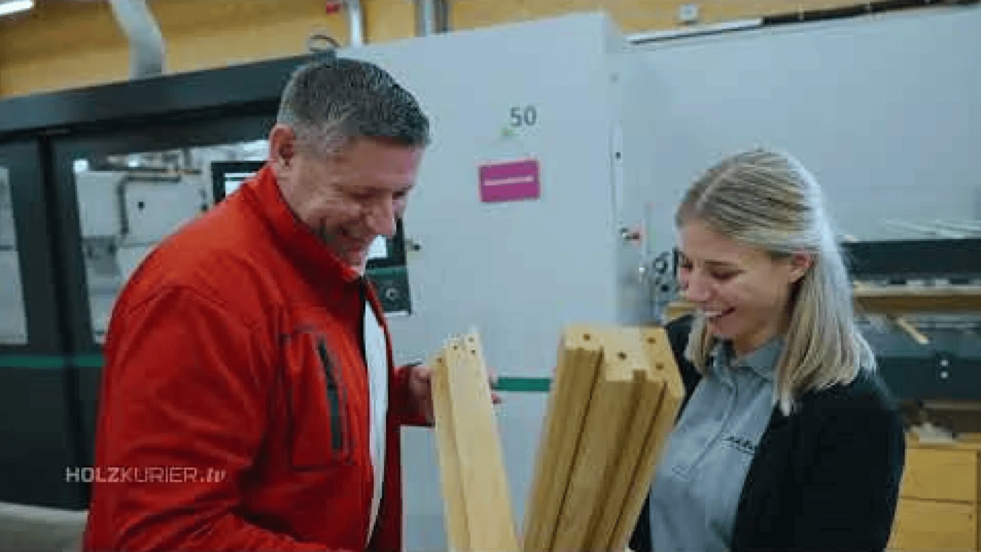 Ein Mann in einer roten Jacke und eine Frau in einem grauen Hemd lächeln, während sie in einer Werkstatt oder Fabrik Holzbretter mit Bohrlöchern untersuchen. Im Hintergrund sind Maschinen zu sehen.