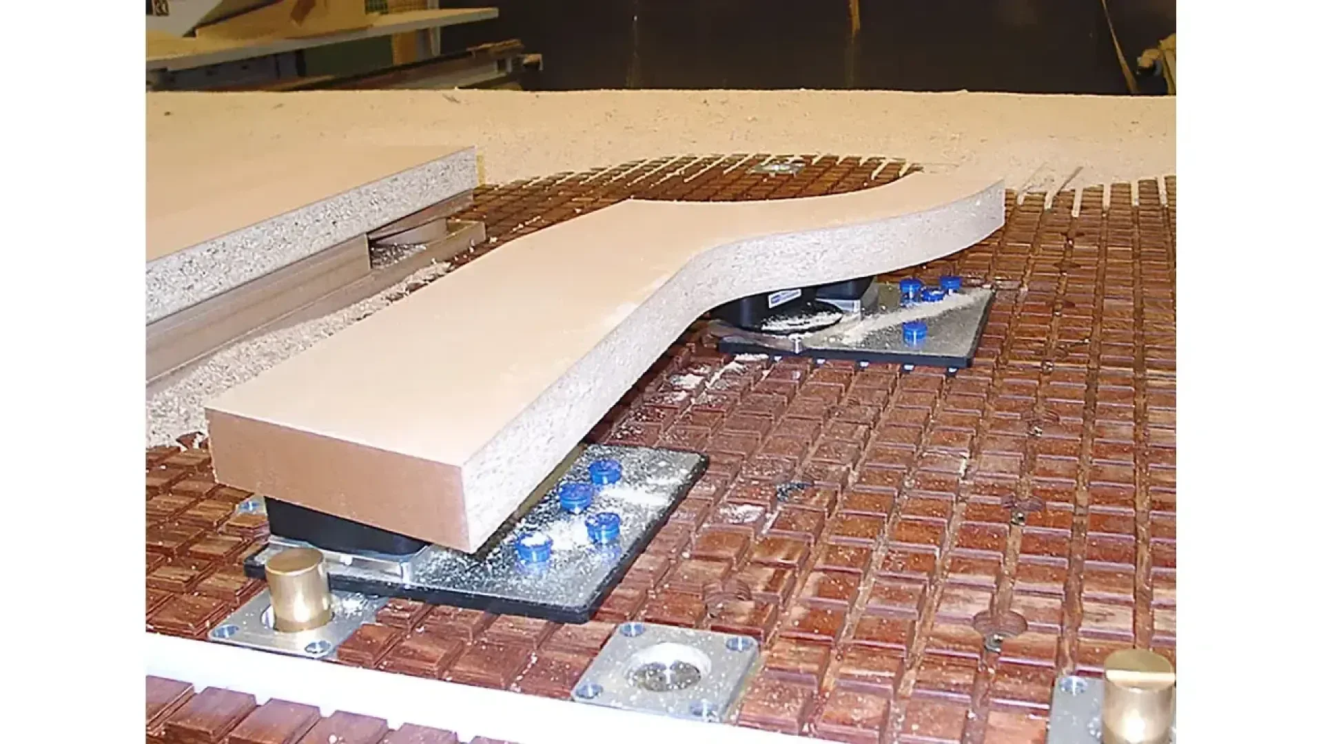 Une machine CNC maintient une planche de bois courbée en place à l'aide de pinces aspirantes sur une table de travail quadrillée, entourée de poussière de bois, au cours d'un processus d'usinage du bois.
