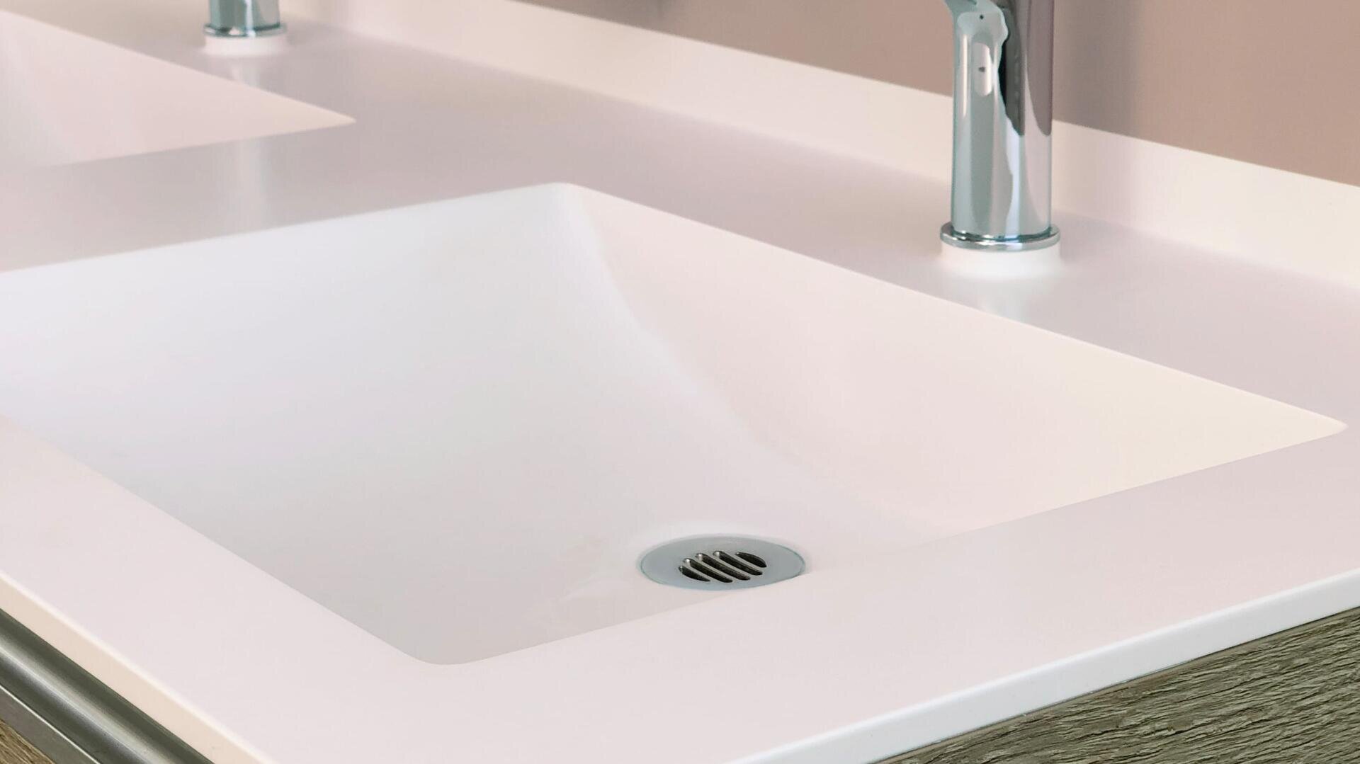 Nahaufnahme eines modernen, rechteckigen, weißen Waschbeckens mit silbernem Wasserhahn und Abfluss, eingefasst in eine glatte Arbeitsplatte. Das Bild zeigt einen Teil eines anderen Waschbeckens im Hintergrund.