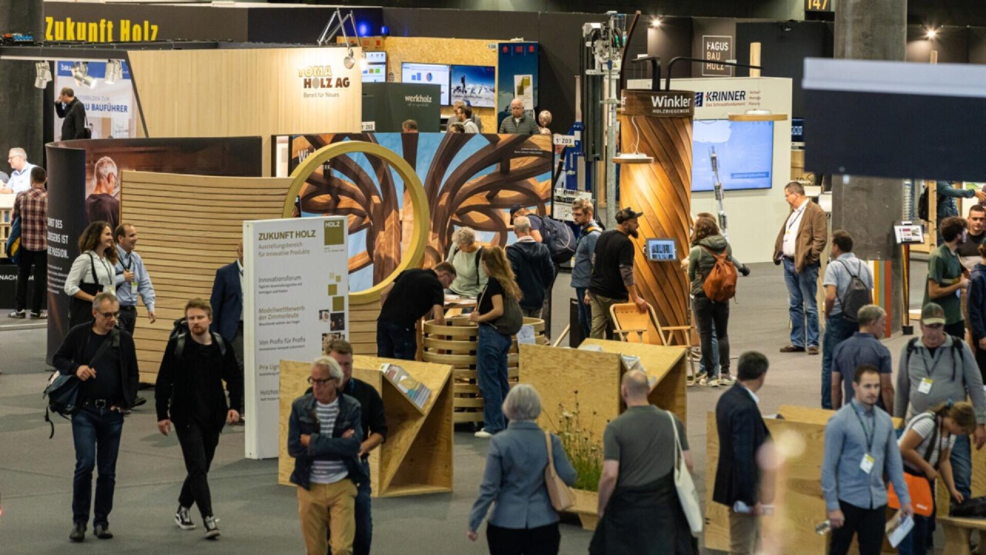 Un hall d'exposition animé où les gens marchent et interagissent autour de structures en bois et de stands présentant des produits en bois et des matériaux de construction. Des panneaux de signalisation et d'information sont visibles dans tout l'espace.