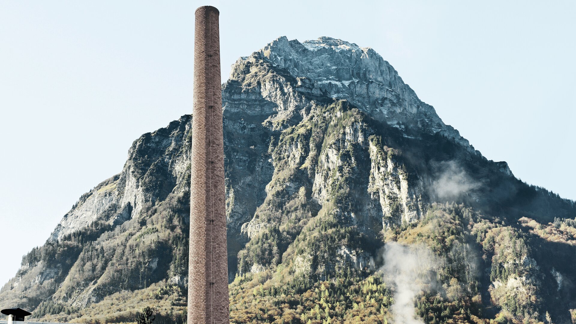 Ein hoher, schmaler Schornstein aus Ziegelsteinen steht vor einem zerklüfteten, bewaldeten Berg mit felsigen Klippen und einer Schneedecke auf seinem Gipfel. Dampf- oder Rauchschwaden steigen in der Nähe der Basis auf. Der Himmel ist klar.