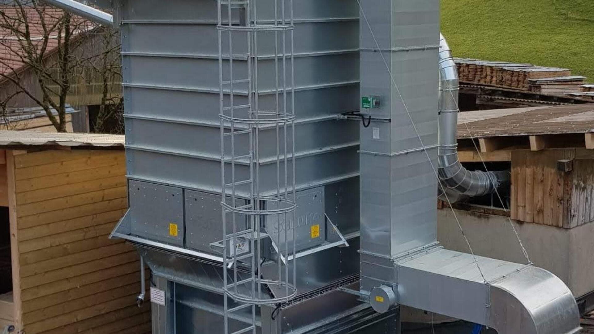 Eine große industrielle Metallmaschine mit Lüftungsschächten und einer seitlich angebrachten Leiter, die zwischen Holzgebäuden steht, mit einem grasbewachsenen Hügel im Hintergrund.