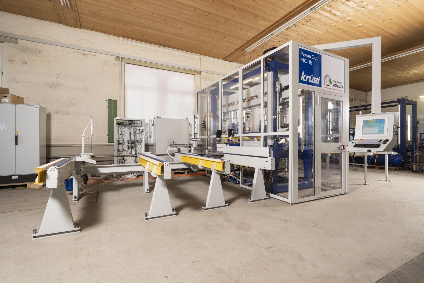 Une machine à bois industrielle moderne avec un système de convoyage, une enceinte de sécurité et un panneau de commande informatique, située dans un atelier spacieux avec un plafond en bois et un sol en béton.