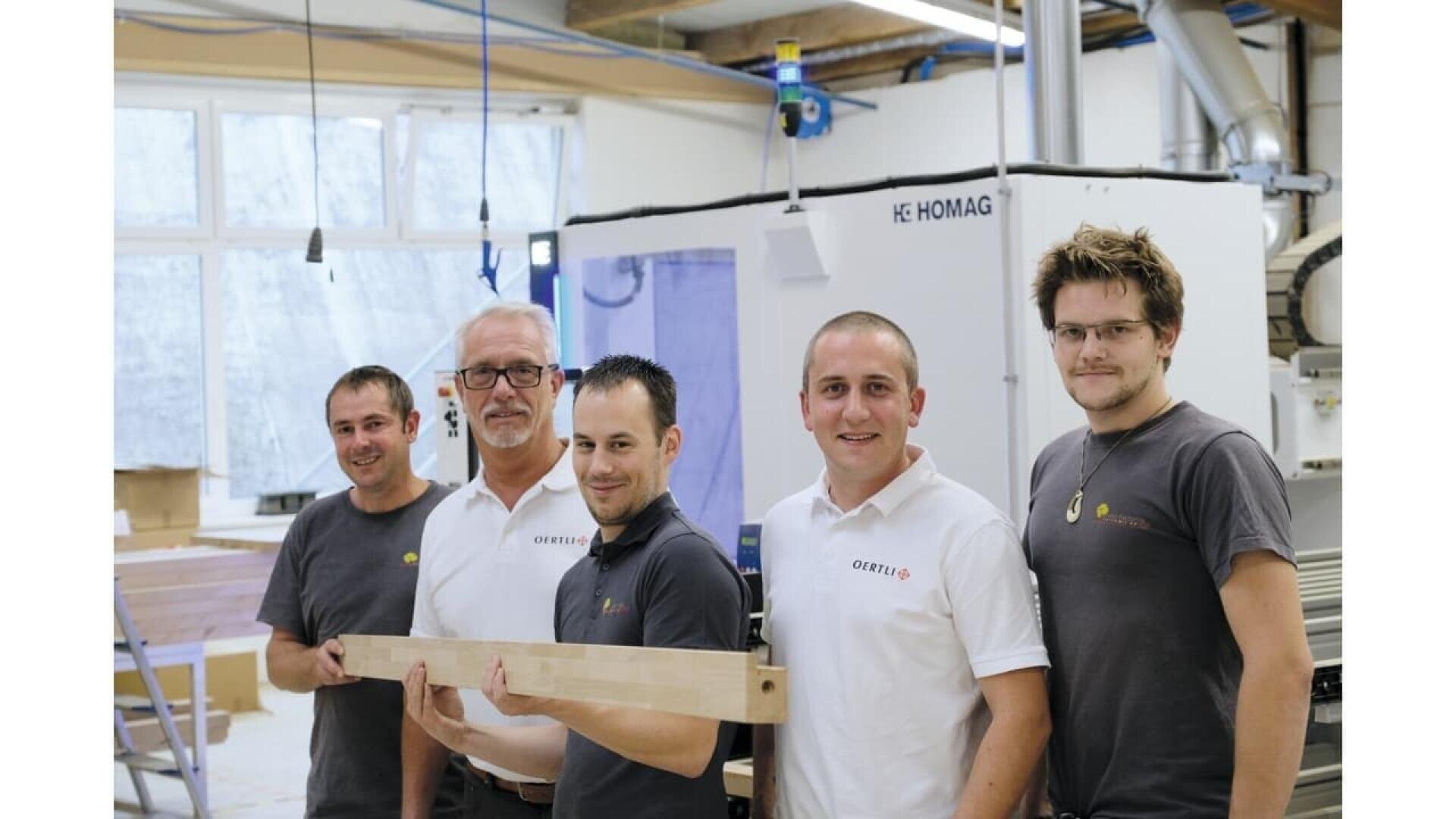 Cinq hommes se tiennent à l'intérieur d'un atelier et sourient à la caméra. Trois d'entre eux portent des chemises blanches et deux des chemises sombres. Ils tiennent une longue planche de bois et des machines et des outils sont visibles à l'arrière-plan.