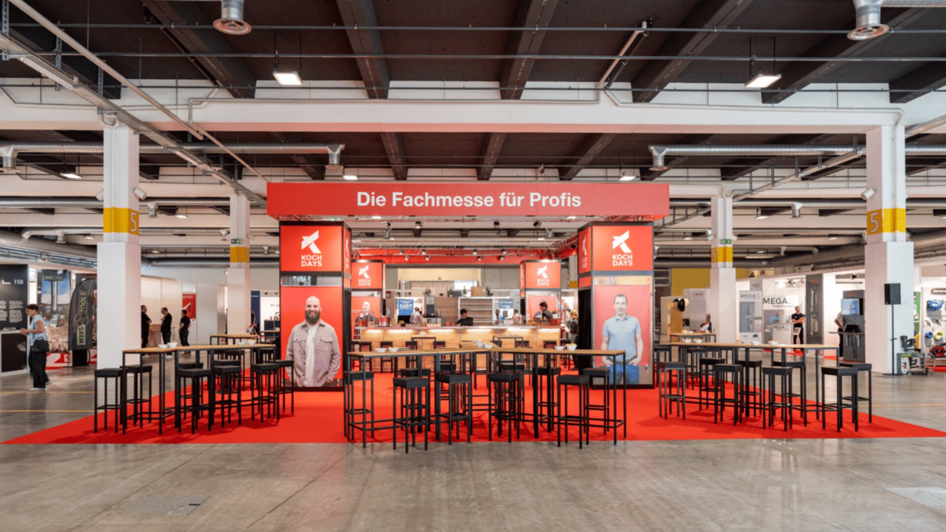 Ein Messestand mit rotem Teppich, Stehtischen, Hockern und der Aufschrift Die Fachmesse für Profis. In einer geräumigen Halle sind große Plakate mit Menschen und verschiedenen Messeständen zu sehen.