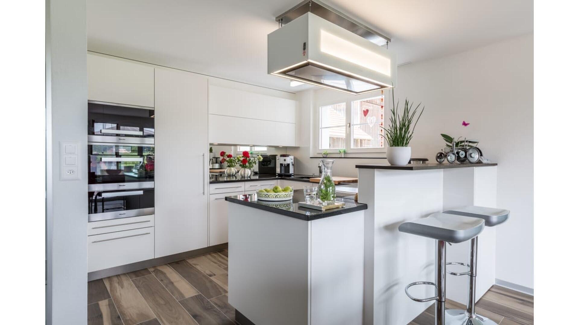 Cuisine moderne blanche avec appareils électroménagers intégrés, îlot avec bouteille d'eau en verre et verres, tabourets de bar, plantes en pot et sol en bois, éclairée par la lumière naturelle d'une fenêtre.