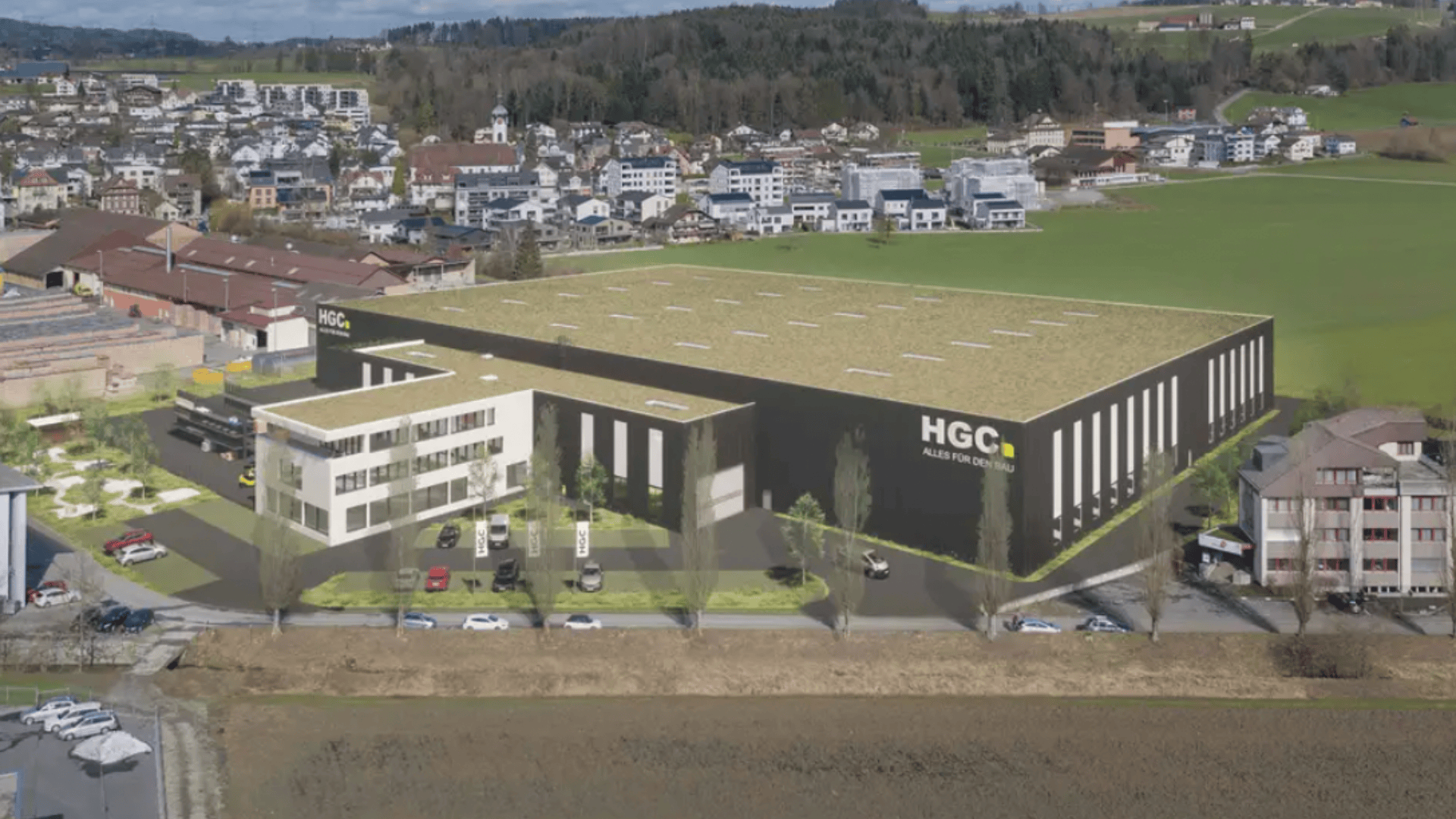 Un grand bâtiment industriel étiqueté HGC avec des bureaux adjacents, entouré de voitures en stationnement, de champs verts et d'immeubles résidentiels en arrière-plan, sous un ciel partiellement nuageux.