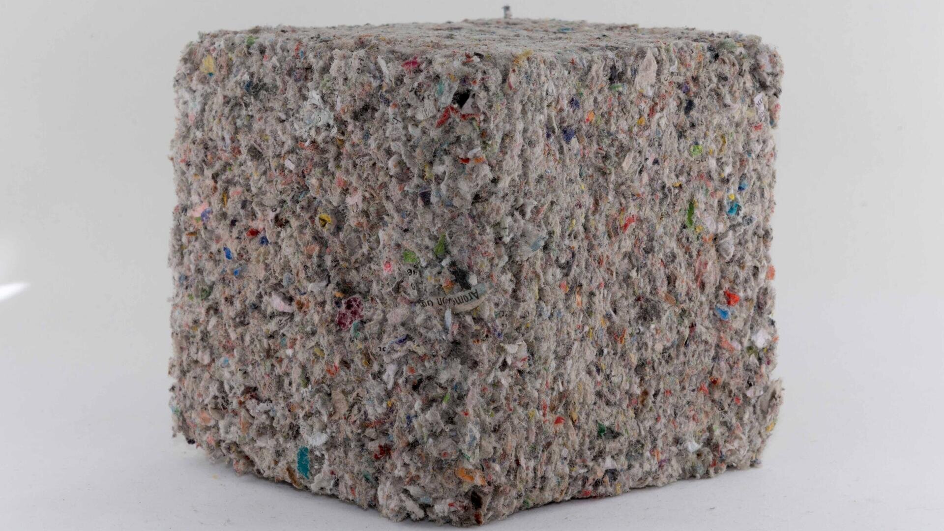 Ein dichter, gräulicher Würfel aus gepressten Recyclingfasern und Flecken aus farbigen Materialien, die wie geschreddertes und gepresstes Papier oder Textilabfälle aussehen, hebt sich von einem einfarbig weißen Hintergrund ab.