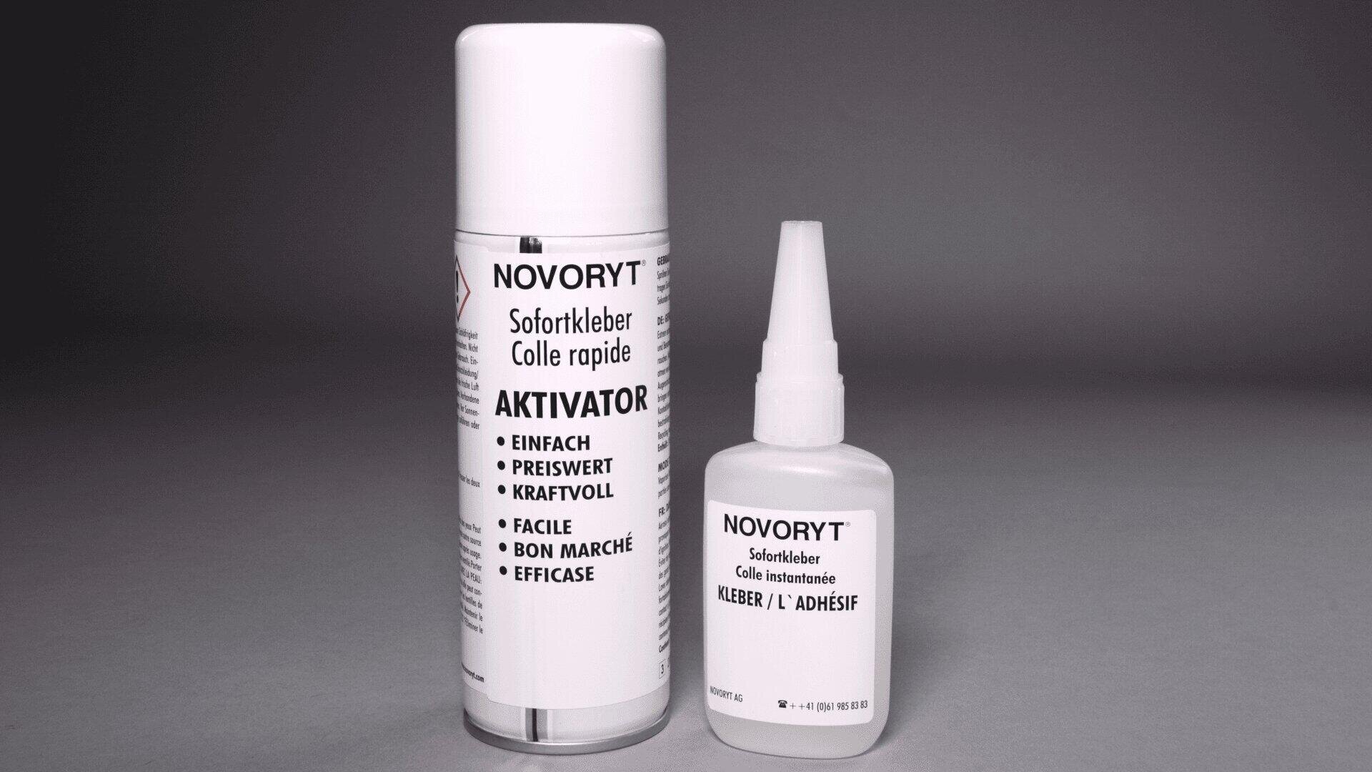 Zwei Klebstoffprodukte von Novoryt sind ausgestellt: eine große Sprühdose mit der Aufschrift Aktivator und eine kleine weiße Flasche mit der Aufschrift Sofortkleber. Beide sind mit mehrsprachigem Text versehen, der ihre Verwendung und Eigenschaften beschreibt.
