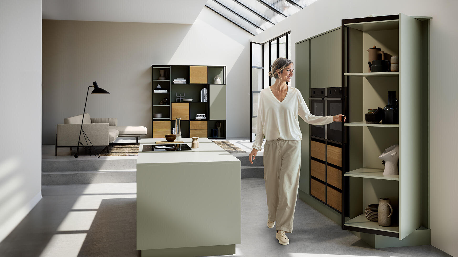 Eine Frau in heller Kleidung öffnet einen hohen, grünen Vorratsschrank in einer modernen, minimalistischen Küche mit einer Insel, offenen Regalen und großen Oberlichtern, die Sonnenlicht hereinlassen.