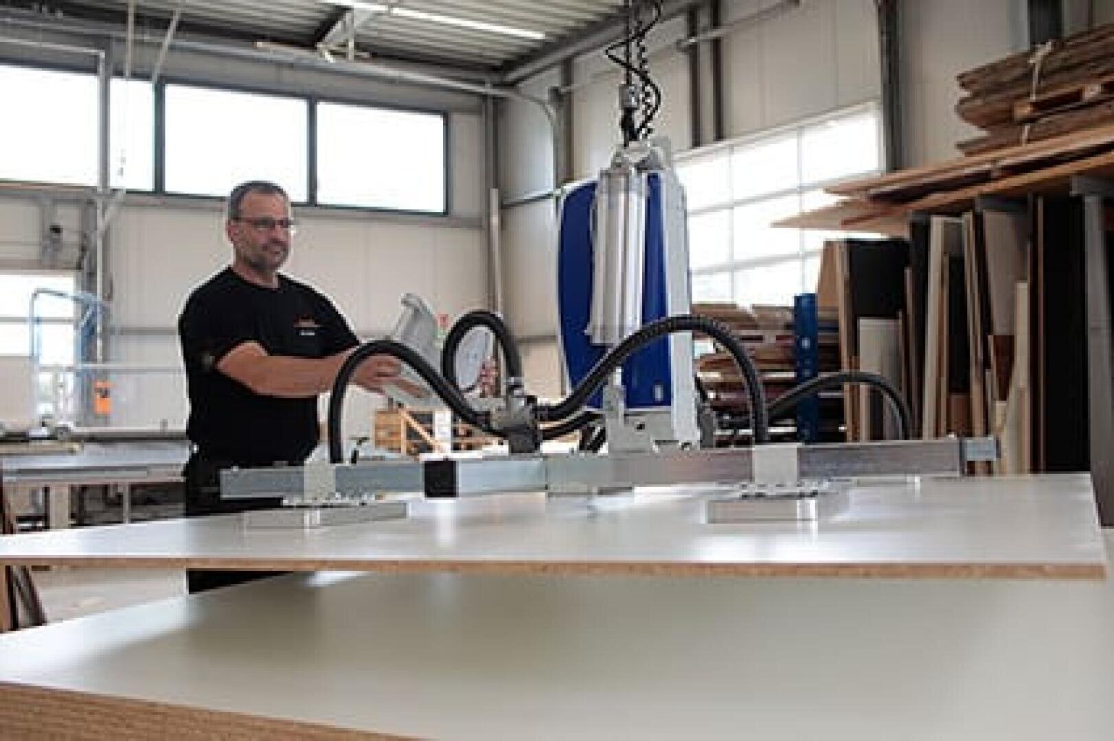 Ein Mann bedient eine Maschine in einer Werkstatt, wobei er eine mechanische Vorrichtung mit Saugnäpfen verwendet, um eine große Materialplatte auf eine Arbeitsfläche zu heben und zu bewegen. In der Werkstatt befinden sich Holzstapel und Industrieanlagen.