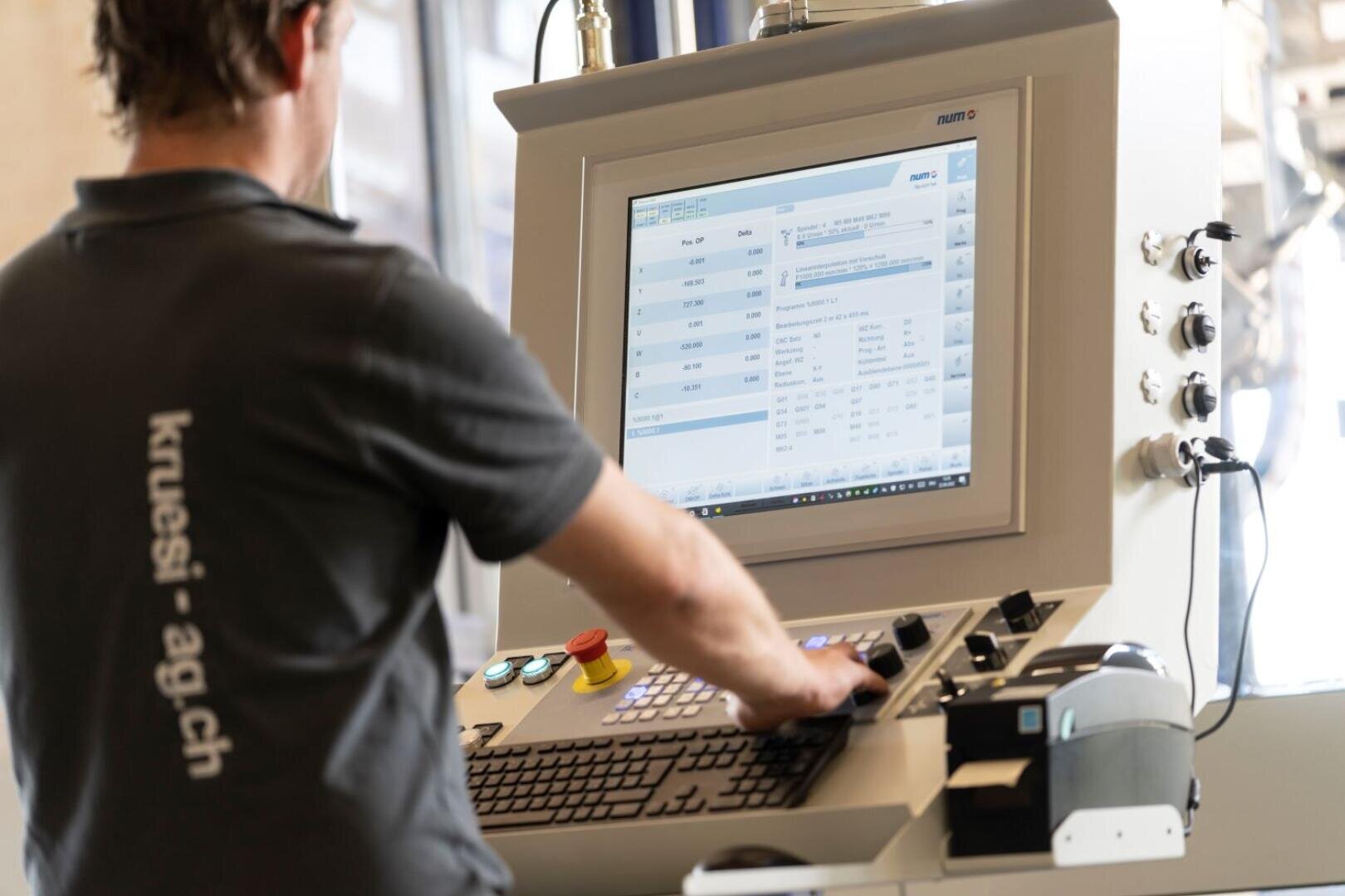 Eine Person bedient ein Touchscreen-Bedienfeld mit verschiedenen Knöpfen und einer Tastatur. Sie trägt ein dunkles Hemd mit dem Aufdruck kruesi-ag.ch auf dem Rücken. Auf dem Bildschirm werden technische Daten und Listen angezeigt.