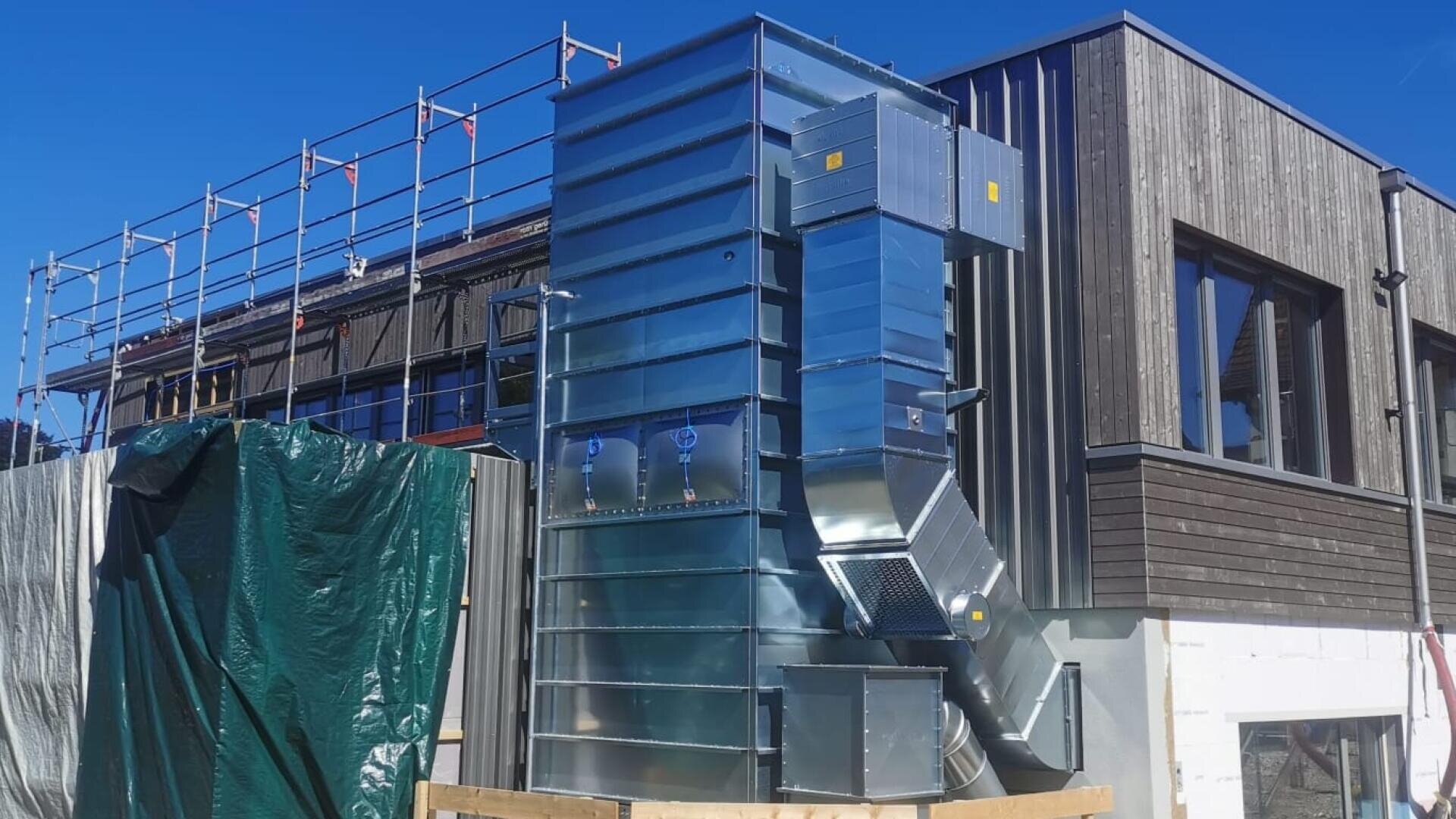 Große metallische HVAC-Einheit, die vor einem modernen, im Bau befindlichen Gebäude installiert ist, mit Gerüsten und hölzernen Außenwänden, die unter einem klaren blauen Himmel sichtbar sind.