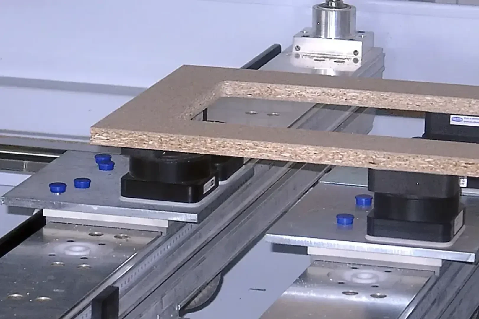 Nahaufnahme einer CNC-Maschine, die ein rechteckiges Stück Spanplatte mit einer Aussparung in der Mitte hält, mit Metallschienen, Klemmen und mechanischen Komponenten.