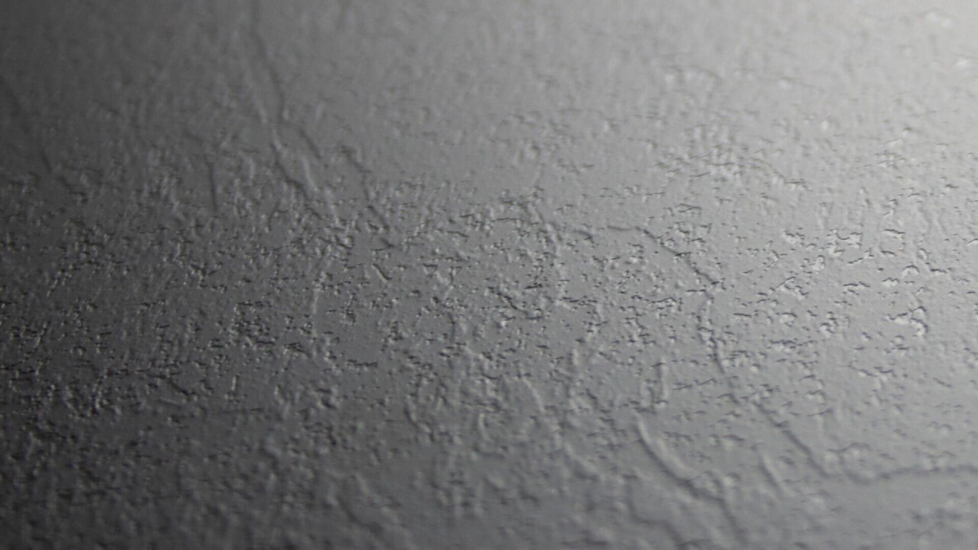Gros plan sur une surface grise texturée, probablement un mur peint ou enduit, avec de subtils motifs irréguliers et une finition mate. L'éclairage met en évidence certaines zones en relief, créant de légères ombres et de la profondeur.