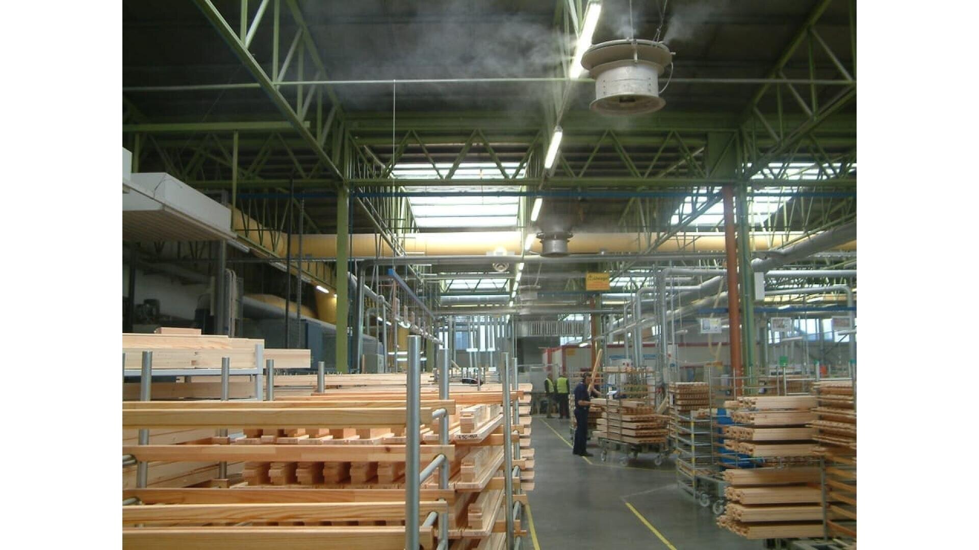 Intérieur d'une grande usine avec de hauts plafonds, des poutres métalliques et un éclairage industriel. Des piles de planches et de pièces en bois sont disposées sur des étagères et des chariots. Quelques ouvriers sont visibles à l'arrière-plan. Un ventilateur émet de la brume depuis le plafond.