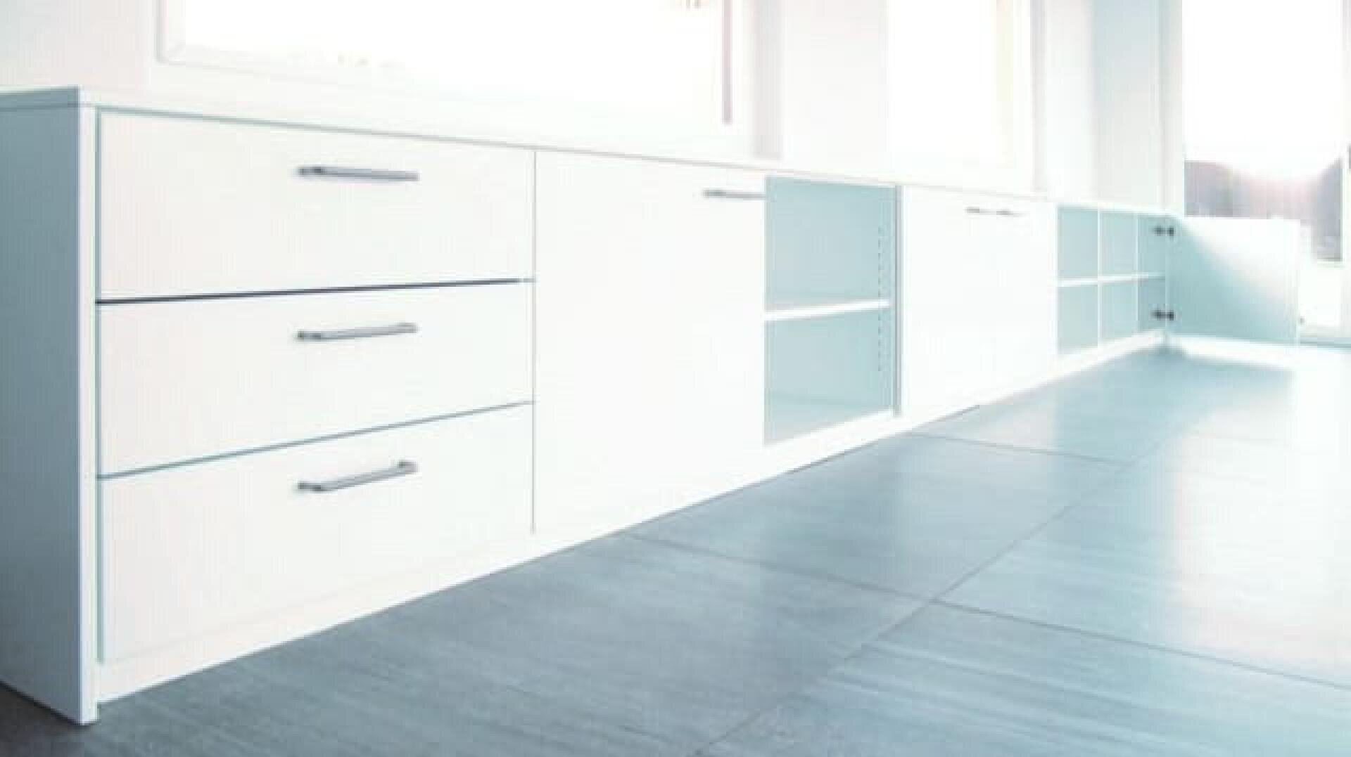 Ein minimalistischer, moderner weißer Schrank mit Schubladen, Regalen und Türen steht an einer Wand in einem hellen Raum mit großen Fenstern und grauem Kachelboden.