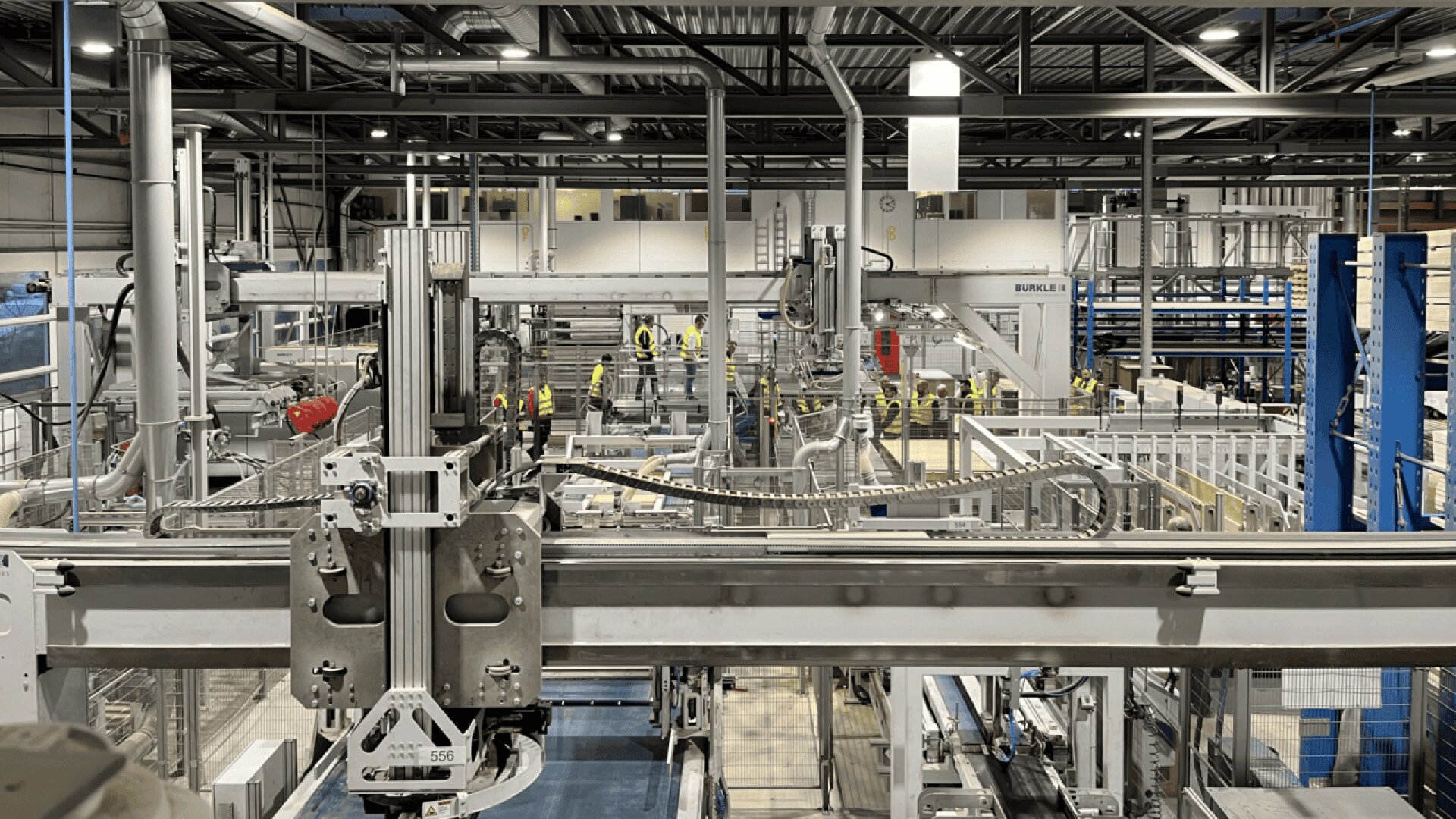 L'intérieur d'une grande usine moderne avec des machines industrielles, des tapis roulants et plusieurs personnes portant des gilets de haute visibilité et des casques de sécurité, ce qui indique un environnement de fabrication organisé et actif.