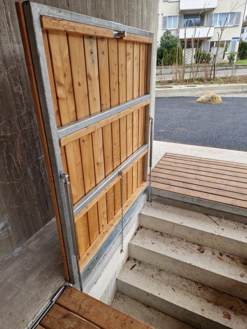 Ein Holztor mit einem Metallrahmen versperrt eine Treppe, wahrscheinlich um den Zugang zu verhindern oder aus Sicherheitsgründen. Das Tor ist teilweise geöffnet und gibt den Blick auf Betonstufen und einen Holzsteg frei.
