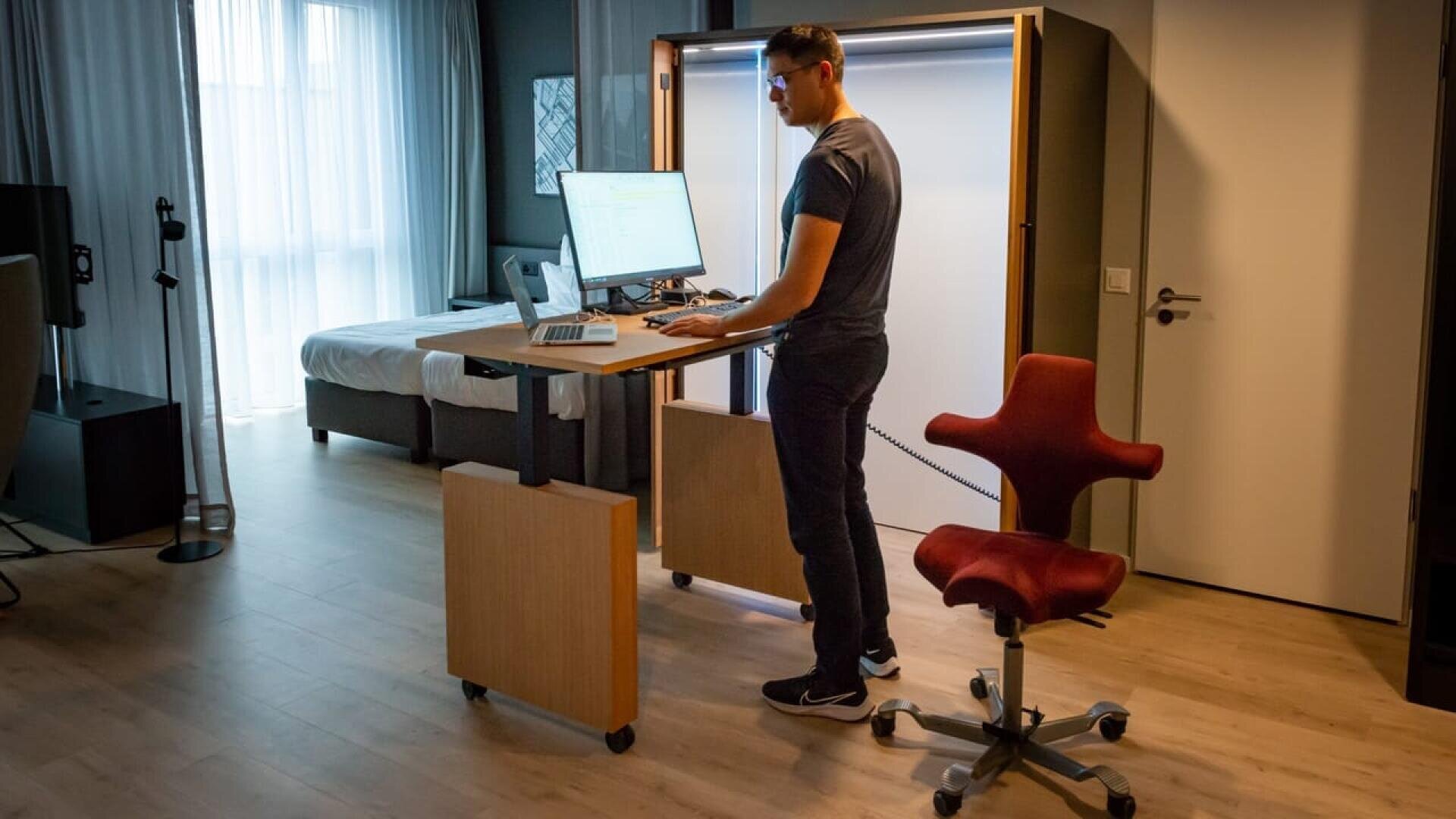 Un homme se tient à un bureau réglable en hauteur avec un ordinateur dans un bureau à domicile moderne et bien éclairé. Une chaise ergonomique rouge se trouve à proximité et un lit est visible à l'arrière-plan.