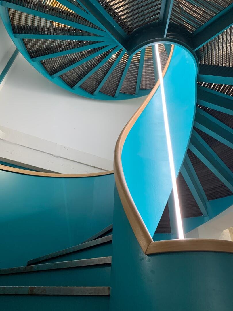 Spindeltreppe mit hellblauen Wänden, hellem Holzhandlauf und einem weißen vertikalen Lichtstreifen entlang der Kurve, von unten gesehen mit Blick nach oben zur Decke.
