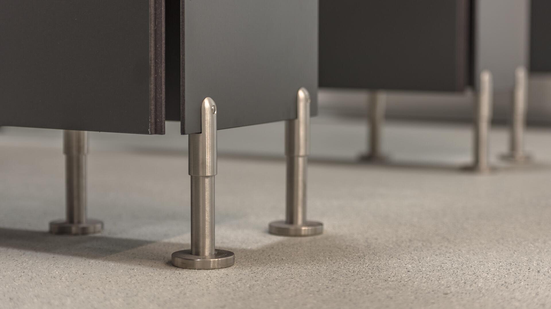 Nahaufnahme von Metallstützen, die an der Unterseite von dunklen Trennwänden für Toilettenkabinen befestigt sind und auf einem hellen, gesprenkelten Boden stehen. Das Bild konzentriert sich auf die Details der Stützen und des Bodenbelags.