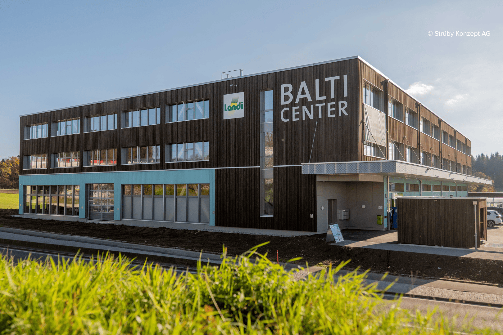 Ein modernes, dreistöckiges Gebäude mit der Aufschrift BALTI CENTER, Holzvertäfelung und großen Fenstern. An der Fassade prangt ein Landi-Schild, im Vordergrund grünes Gras und klarer Himmel.